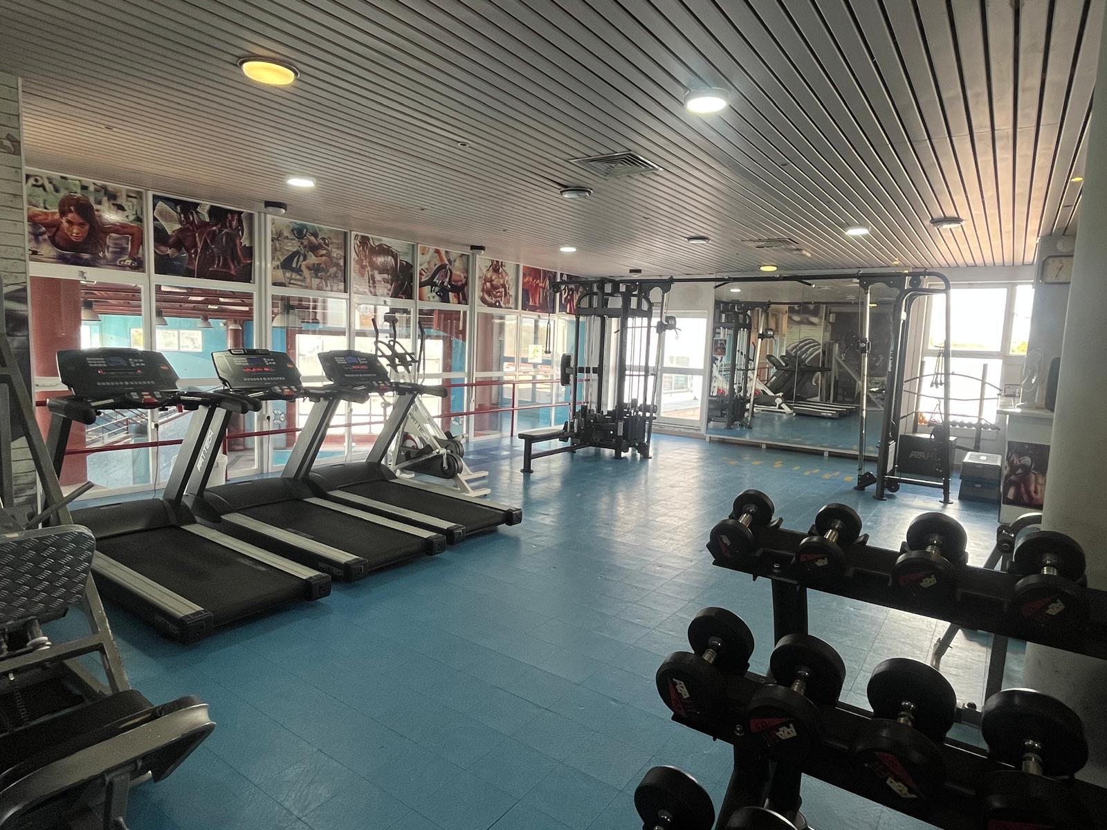 Sala de ginásio com três passadeiras, máquinas de musculação e halteres dispostos em suportes, piso azul e janelas grandes.
